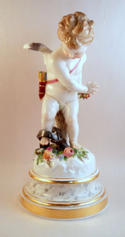 Meissen Figure of Cupid Trapped: Meissen porcelain figure of Cupid caught in a bear trap. The figure holds a small bouquet of flowers in his hand and has his quiver full of arrows across his back. A large bear trap ensnares Cupid's a