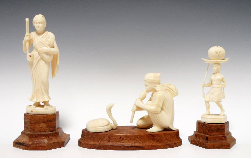 Indian Ivory Carved Figures (3): Group of three Indian ivory carved figures. One figure is of a snake charmer, the second is of a man with a basket on his head, and the third is of a woman holding a baby. Each piece rests on a wood b