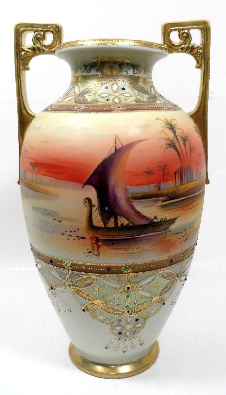 Nippon Hand Painted Two-Handled Vase: Nippon hand painted two-handled vase. Warm tones and elaborate gilt enamel work decorate this tall vase. A Middle Eastern oasis is painted onto the middle of the piece. The two handles are painted gol