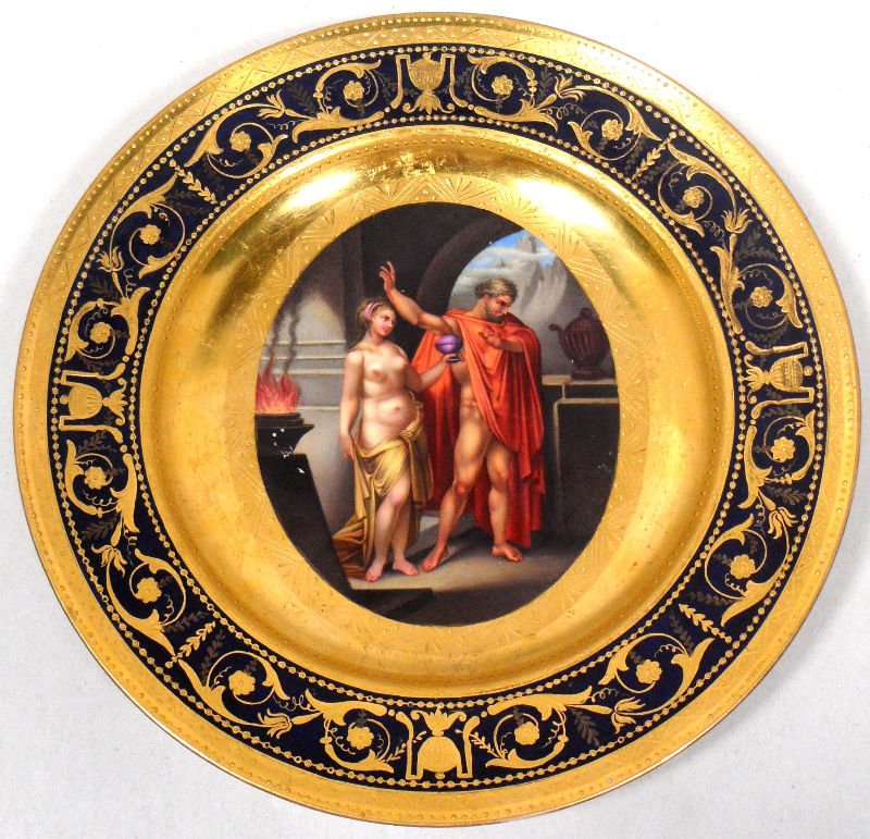 Royal Vienna Painted Porcelain Plate: Royal Vienna painted porcelain plate. The center of the plate depicts a scene of a woman offering a man a chalice. The piece is heavily gilt with intricate Grecian patterns and features a cobalt blue 
