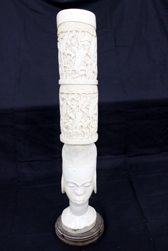 African Carved Figural Ivory Tusk: African Carved Figural Ivory Tusk. Depicting a woman's face with a hat of figures. Mounted on wooden base. Weight - 12.29 pounds. Measures - 24 3/4" high x 8" wide. THIS LOT IS THIRD QUARTER 20TH CENT