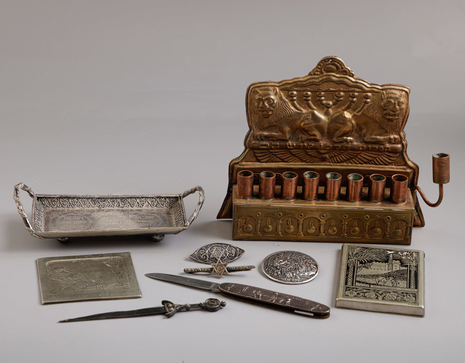 Bezalel Items: Lot of Bezalel items, made of various materials including silver, copper, and metal. Comprising a Hanukkah lamp, plaque, box, bottle opener, tray, pins, clasp, and pocket knife. Decorated with filigre