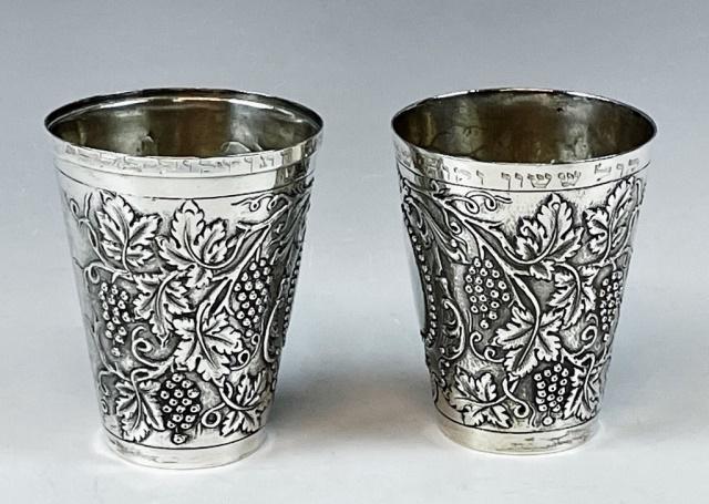 19th C Silver Repousse Kiddush Cups, Germany (2pc): Pair of .800 Silver Kiddush Cups, Germany. Crafted in silver with repousse grape and vine decoration with hand chased embellishment. Rims carry chased Hebrew inscription. Excellent condition, measures