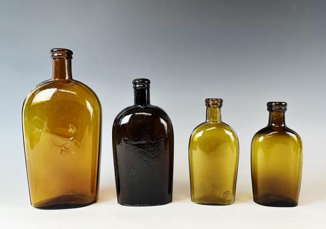 19th C Whiskey Bottles Baltimore, Whitney (4pc): Collection of Four 19th C. Blown Glass Whiskey Flasks: (1) Amber flask, base signed "Whitney Glass Works" (1) Amber colored flask embossed with anchor, Baltimore Glassworks (1) Dark glass strap sided