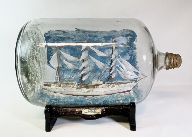 Folk Art Ship Model in Bottle, USCG Eagle: Folk Art Ship Model in Bottle, USCG Eagle. Large clear glass bottle with plastic model of the USCG Ship "Eagle". Bottle on wooden stand with plaque. Good condition, measures: 13.5" H x 20.5" L. Model 