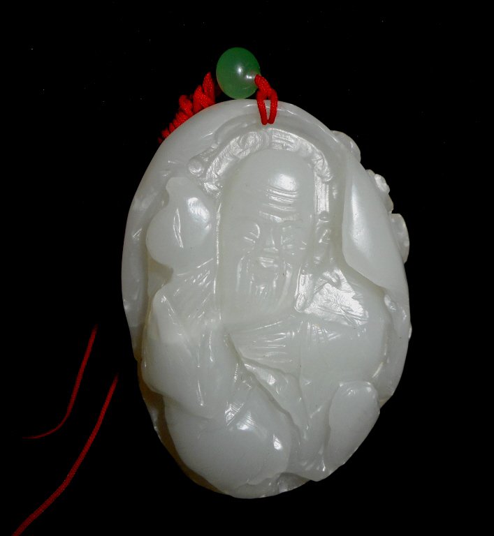 Jade Shoulao Pendant: White jade pendant suspended on red silk cord with small green jade bead depicting Shoulao, God of Longevity holding a peach carved in bas-relief, surmounted by a bat. Smooth polished surface with inc