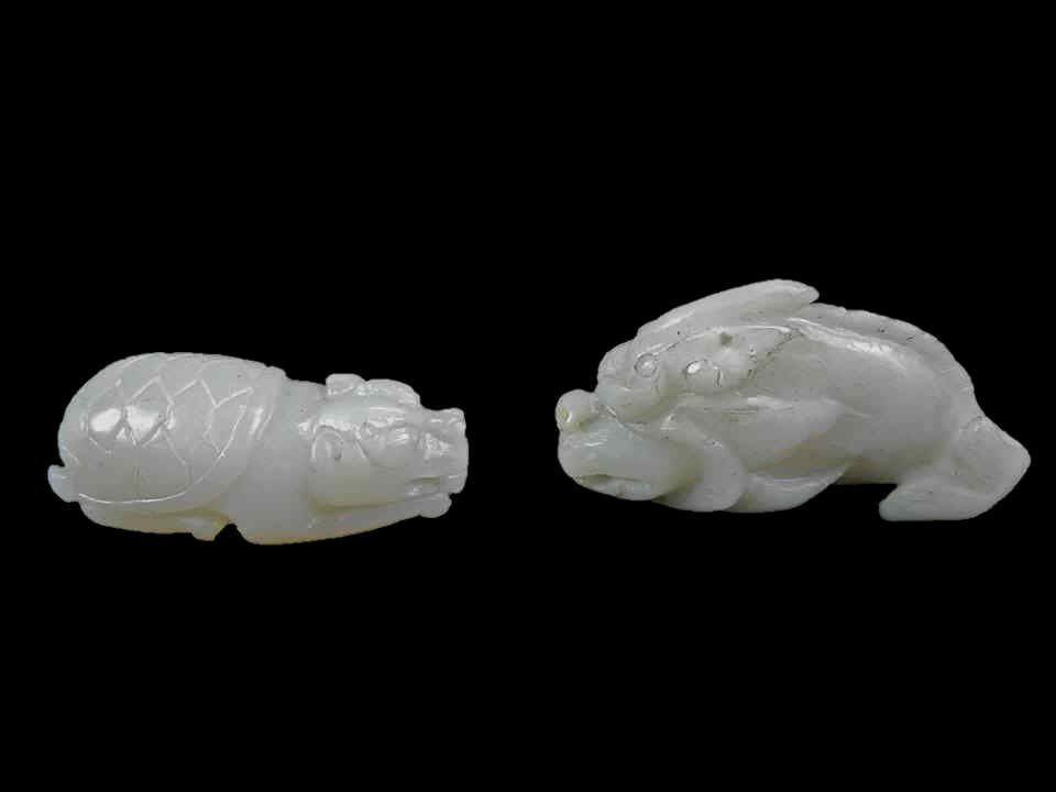 Two Jade Pendants of Mythological Beasts: Two white jade pendants in the form of mythological beasts, each suspended from a red silk cord, having smooth polished surfaces with incised details. One pendant is fashioned in the form of a recumbe