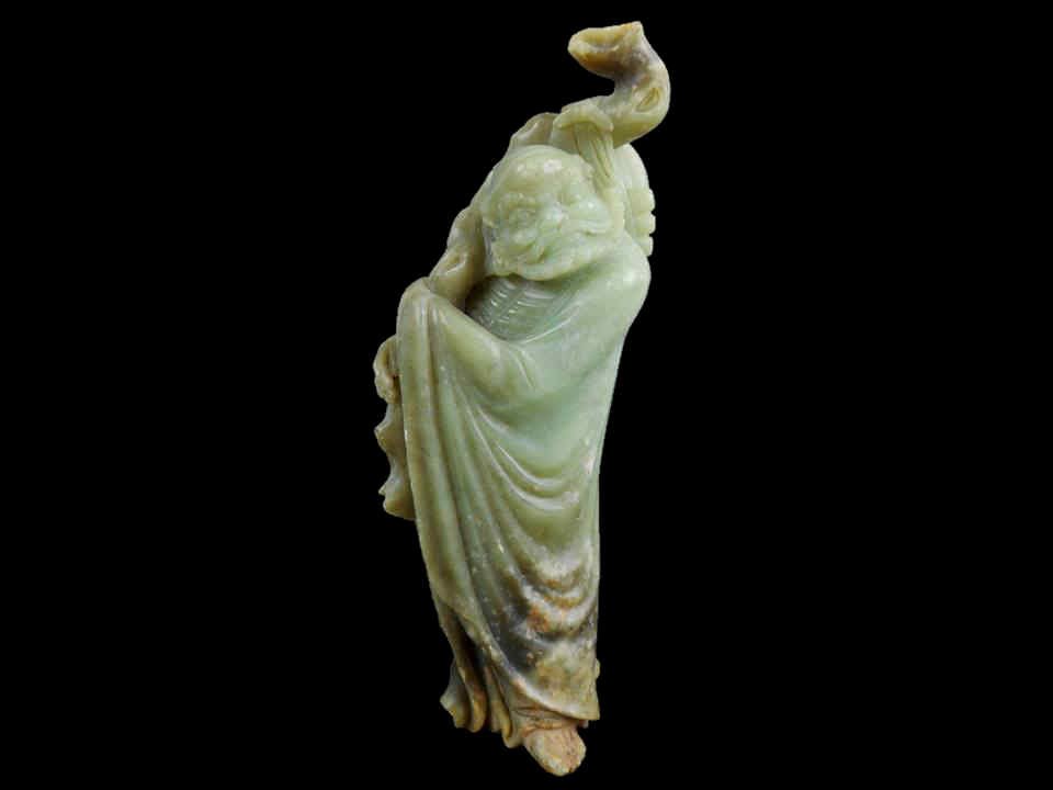 Jade Figure: Green carved jade figure of a man in robes, holding a staff with a Bi affixed to his back. Smooth surface with incised features, tan, russet and brown suffusion, calcification and erosion. 10"H