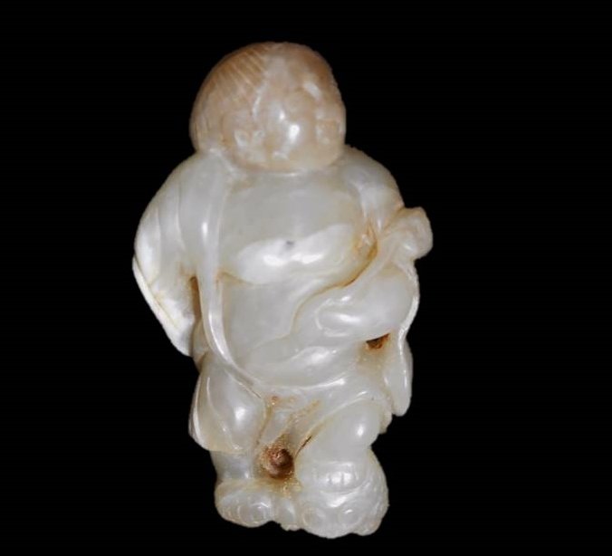 Jade Figure: Grey-green jade carving partially robed figure holding a lotus frond behind his back, a string of coins and a three-toed frog underfoot. Smooth surface with incised details and areas of brown and whit