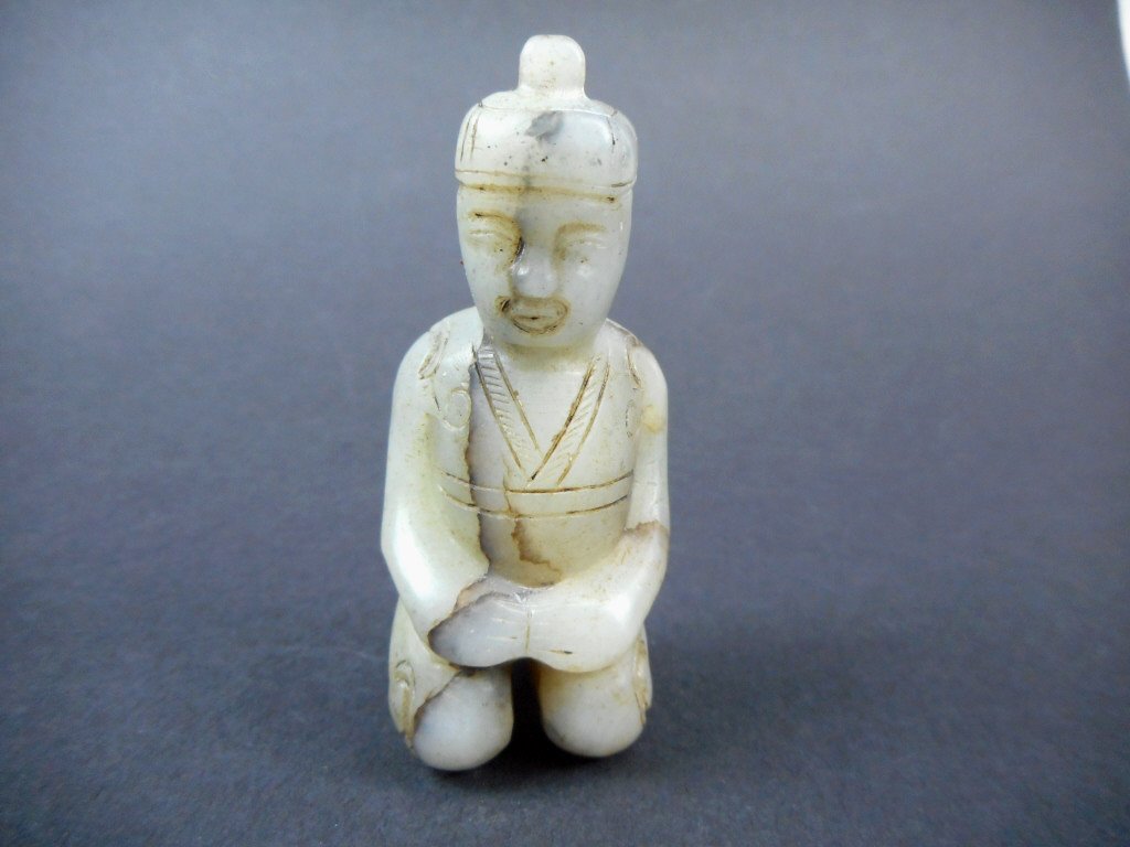 Jade Pendant: Grey-white pendant in the form of a kneeling soldier figure in robes. Dark brown and black suffusion and veining. Darker area at feet, suggestive of burial. 3"