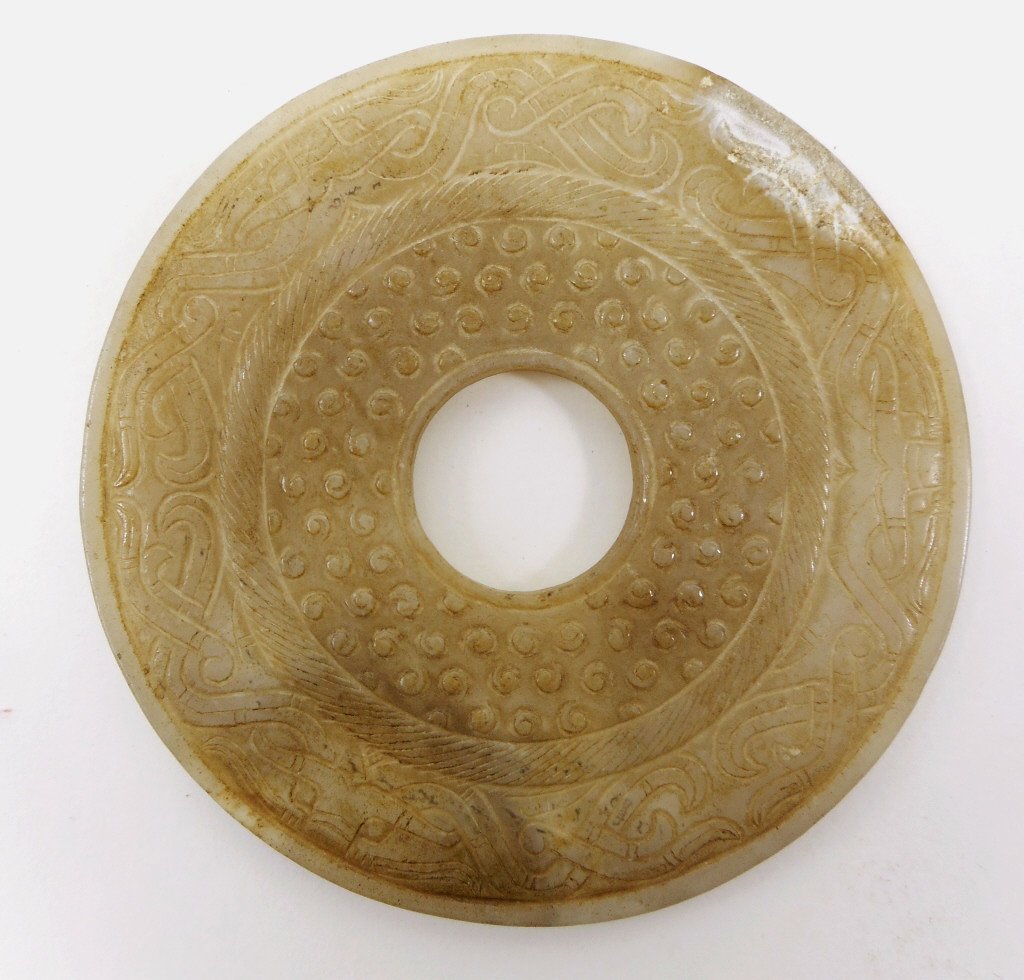 Jade Bi Disc: Small white jade Bi disc with incised rice grain pattern and anthropomorphic mask design. Calcification and dark brown suffusion suggestive of burial. 3"