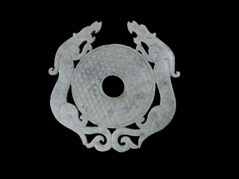 Jade Bi Ornament: Green jade ornament in the form of a Bi with incised rice grain pattern, flanked by two dragons with stylized phoenix head tails. Smooth surface with diffuse white calcification. 6"H x 6" W
