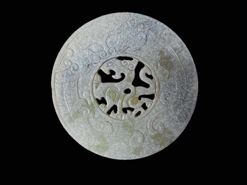 Jade Ornamental Disc: Green jade disc with incised dragon, phoenix and anthropomorphic mask designs carved in bas-relief. Center features openwork rendering of a dragon. Diffuse calcification with darker areas of suffusion
