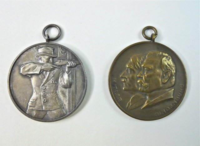 Two German 1930's Schutzen Awards: One in silver depicts shooter with rifle aimed and swastika flag in background. Reverse reads, "20 DEUTSHES BUNDESCHIESEN LEIPZIC 1934" in gothic font. Edge is stamped "SILVER 1000 FEIN." Measures 1.8