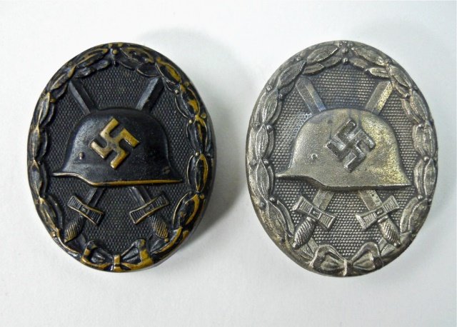 Two WWII German Wound Badges, Black and Silver: Die struck alloy, silver wash with Swastika emblazoned helmet surmounting crossed swords in oak leaf wreath. Reverse with hinged pin and c-form catch marked "92". 1.75", 24.1 g. Black painted brass wi