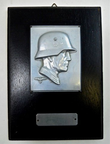 WWII German Military Wall Plaque: Black painted wood with silvered metal profile of German soldier wearing steel helmet with small NSDAP eagle at shoulder signed, "BM" in circle. Small rectangular metal tag beneath blank without perso