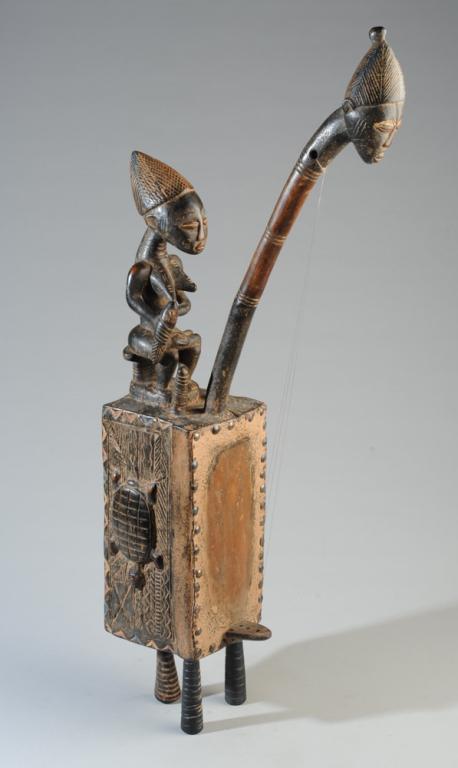 Baule Figural Box Harp: Carved wood stringed instrument with a woman nursing a child on the top, the sides with a female face in relief on one side and a turtle on the other, with animal hide covering the sounding board, thr