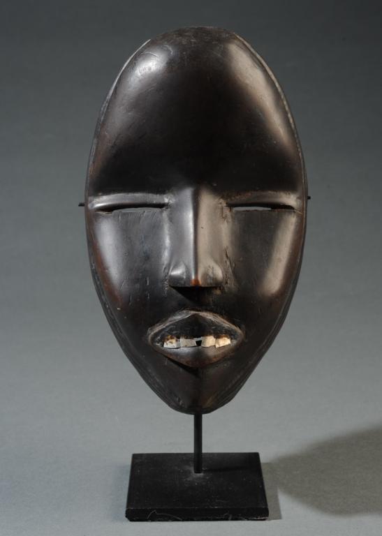 Dan Deangle Mask Metal Teeth: Expressing a peaceful, feminine spirit, with narrow eyes, delicate features, a dark, burnished surface, and enhanced with metal teeth. Mask is very thin-walled, appears to have age. On gallery stand. 
