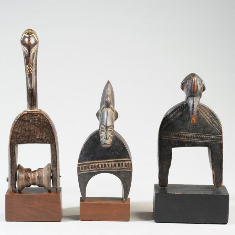 Three African Heddle Pulleys: Probably Senufo. One anthropomorphic pulley, the figure with crested coiffure, coffee bean eyes, facial scarification, the other two pulleys with bird-like finials. On gallery stands. Largest pulley,