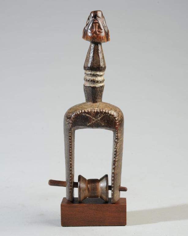 African Figurative Heddle Pulley: Phallic form relief carved with four Baule style masks, three are horned / zoomorphic, the fourth a human face turned upside down. On gallery base.Pulley, approx. 8 3/4" H. Provenance: Estate of Thoma
