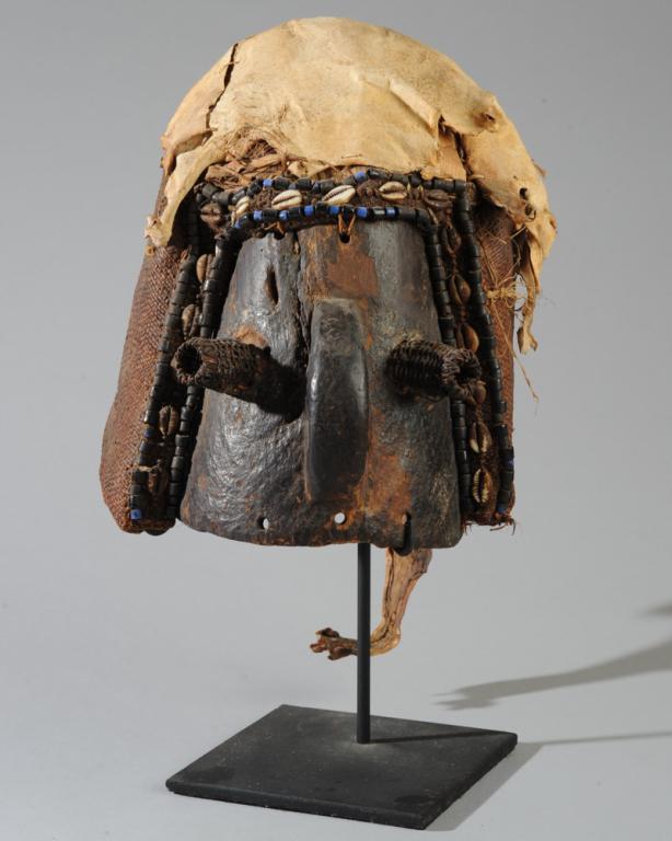 Binji Mask, D. R. Congo: The face wood with wooden nose and protruding basket-weave eyes, the helmet of woven fiber edged with blue and black glass trade beads and shells, the top of the helmet covered with the hide of a smal