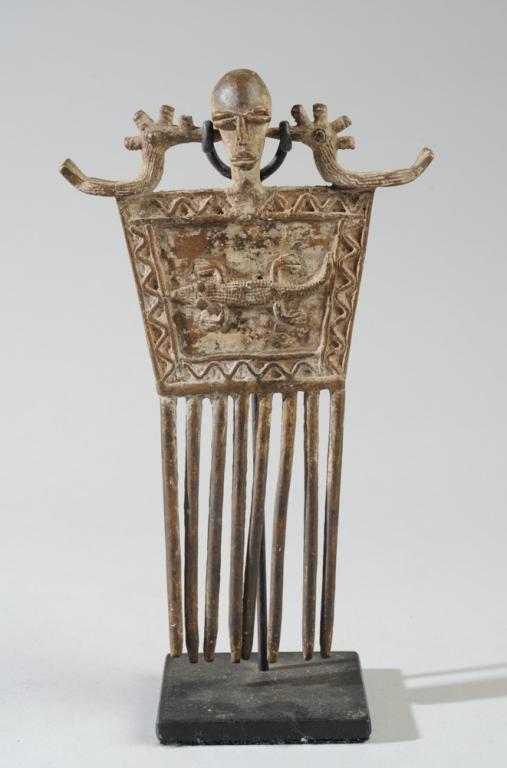 African Bronze Figural Comb: A head flanked by two birds above a square body with a crocodile in relief on one side and what appears to be a large arachnid or beetle on the other. Eight tines. Approx. 7" H. On custom gallery base