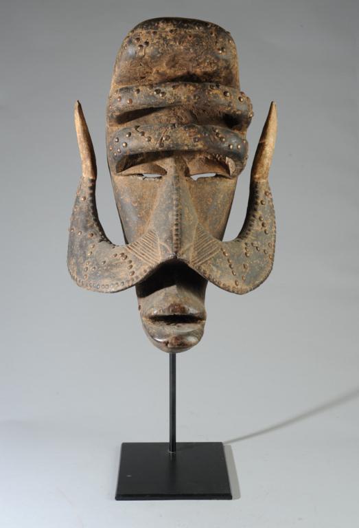 Bete Warrior Mask, Ivory Coast: Large carved wood mask with metal studs, rough encrusted surface to exterior, interior smooth and glossy. Bete people, Ivory Coast. On custom gallery stand. Approx. 21" long. Provenance: Estate of Tho