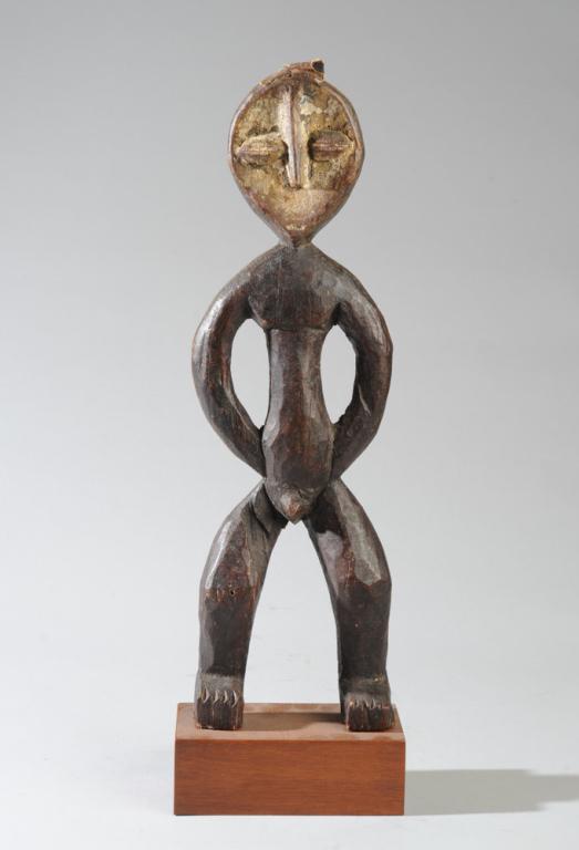 Lega Figure: Carved wood standing male figure with arms akimbo, the concave mask face with applied kaolin pigment. Small piece of animal hide nailed to top of head. On gallery base. Approx. 17 1/2" H, 19 1/2" H wi