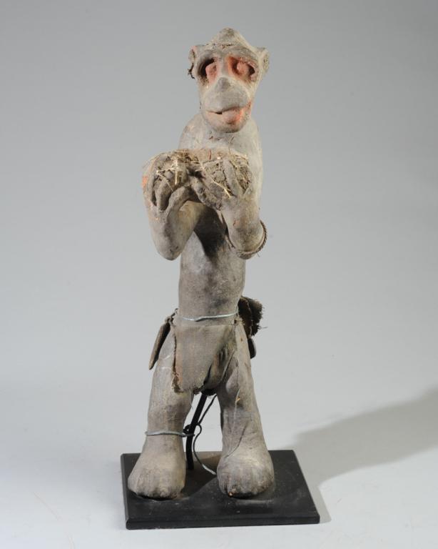 Baule Gbekre Mbra Monkey Figure: Heavy blackened terracotta monkey figure with reddened eyes, carrying a vessel with vestiges of organic material including feathers. Small objects tied to the figure's waist. Chip to one ear. Minor lo