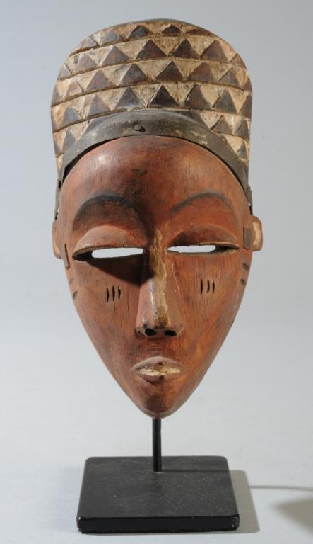 Eastern Pende Mask: Small anthropomorphic mask with delicate heart shaped face with half-closed, heavy lidded eyes under arched brows and with a high coiffure or headdress embellished with relief carved triangles highlig