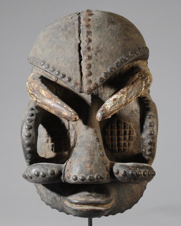 Bete Mask: Intimidating mask of carved wood with many metal studs and crusty surface, having a heavy overhanging brow, large nose, protruding duck-bill lips, and large curving horn-like projections. Bete people,