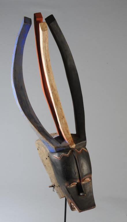 Guro Antelope Mask, Ivory Coast: Black antelope face with half-moon eyes, very human nose, open mouth with pointy white teeth and red tongue, and crowned with long curving red, white and blue horns. Interior with bite bar. On custom
