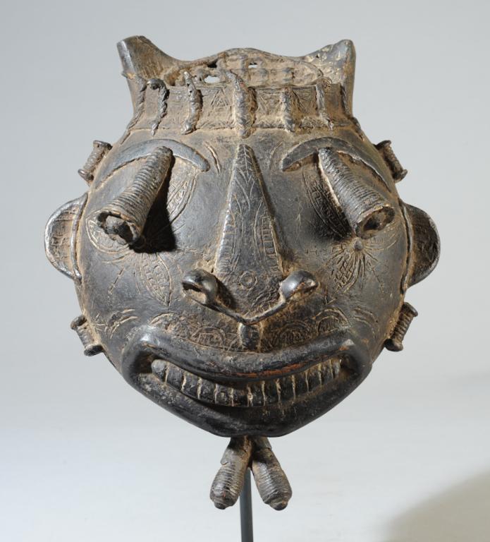 Tikar Bronze Mask: Cast bronze mask with complex coiffure or headdress above the round face, the eyes in conical stalks jutting out beneath arched brows. Below the wide grinning mouth, the form of the eyes is repeated i