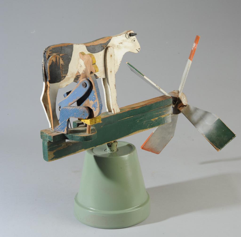 Antique Folk Art Whirligig with Cow: Carved and painted whirligig featuring a bearded farmer in yellow hat and blue coverall, milking a black and white cow when the wind blows. Early 20th C. Minor losses to painted surface. Approx. 17" l