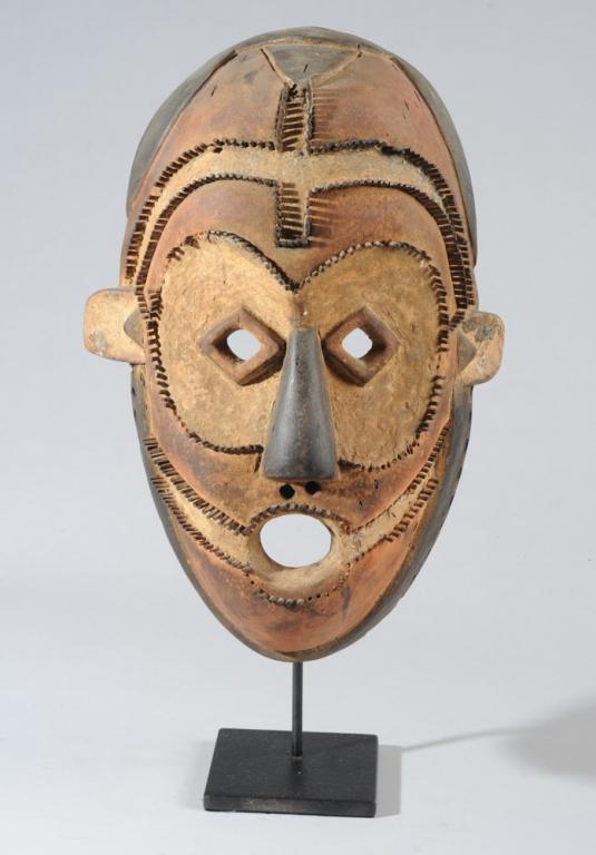 Ngbaka Mask, Democratic Republic of Congo: Carved wood mask with orange, white and black pigment, the graphic elements of the mask outlined with rows of tiny wooden pegs. Small round eyeholes set in relief diamond shapes within large oval slig