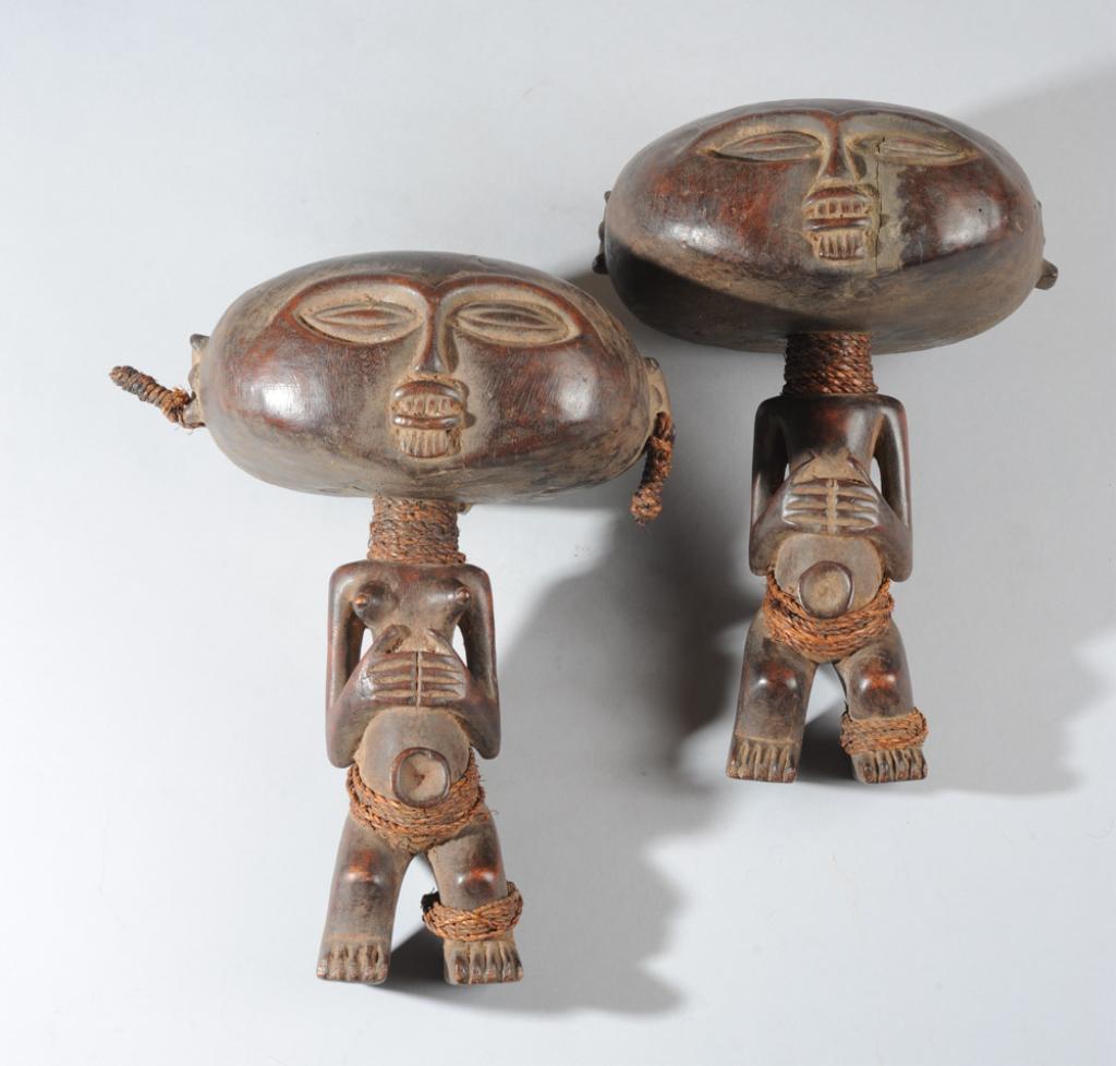 Tabwa Male & Female Figural Rattles: Carved wooden pair with large, smiling, childish heads, posed identically with hands clasped over abdomen above prominent umbilicus, both with twine wrapped at neck, waist and left ankle. The female w