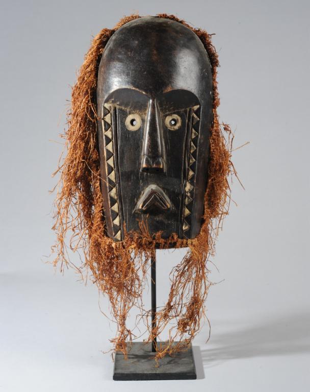 Bamana Mask: Anthropomorphic mask with graphic white on black sawtooth pattern from temple to jaw. Brow, nose, and downturned mouth carved in bold relief. Long raffia fringe. Bamana artist, Mali. Mask, approx. 14"