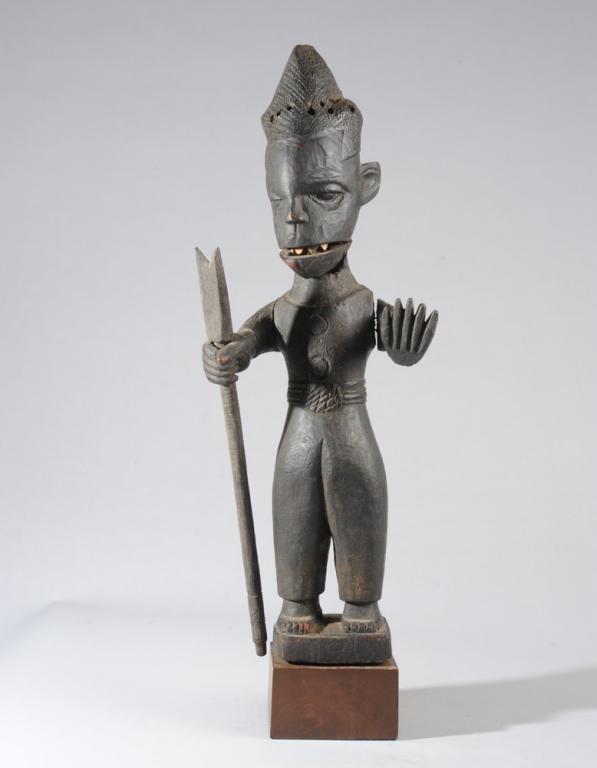 Ibibio Puppet, Nigeria: Carved wooden male figure in official-looking garb, a staff or weapon on one hand, the other raised as if to say Halt! The lower jaw is articulated, revealing sharp white teeth when the mouth is open.