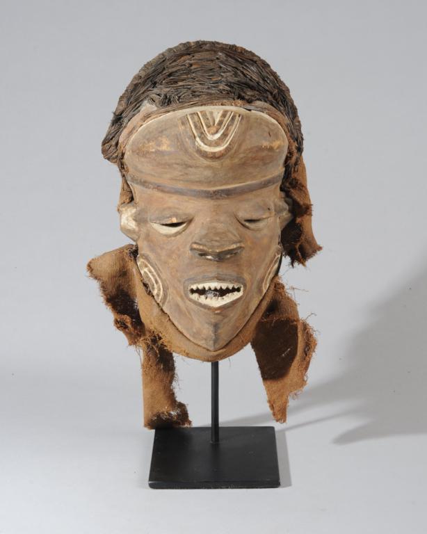 Pende Mbuya Mask: With bulging forehead, relief carved facial markings, lips parted to reveal pointed white teeth. Raffia and fabric headdress and collar. Repaired break to left edge from temple to jaw. On custom galle