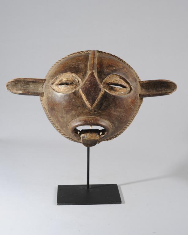 Kwele Mask, Gabon: Round face mask with long horizontal ears, the large coffee bean eyes set within whitened hollows, the cranial ridge extending to diamond shaped nose above open mouth with protruding tongue and animal