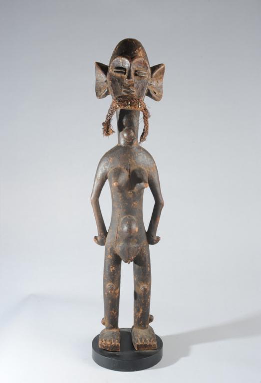 Female Power Figure, Rwanda: Older carved wood figure of a youthful female with domed head and heart shaped face, the simplified features (eyes and lips represented by identical parallel lines in high relief) expressing both conc
