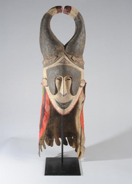 Igbo Helmet Mask, Nigeria: Likely an agbogho mmwo / maiden spirit mask, with tall, red-tipped horn-like coiffure, refined facial features, the lips parted and curved in a smile. The mask retains its marvelous red, yellow, green