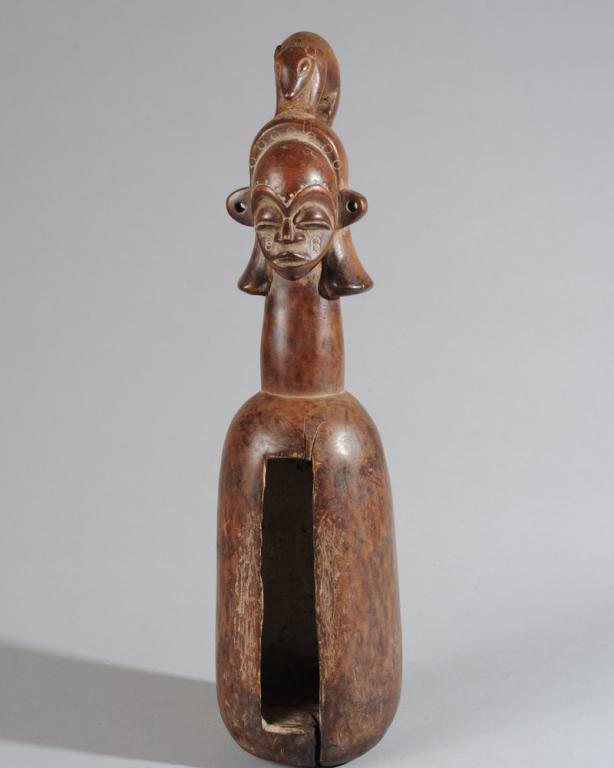 Yaka Slit Drum: Carved wooden slit drum topped with anthropomorphic head with large, closed eyes, arched brows, and with long tailed animal perched atop coiffure. Such drums, called n'koku ngoombu, have various uses 