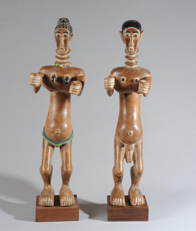 Fante Male and Female Figures, Ghana: Pair of carved figures of light brown wood, the hair, eyes, eyebrows and breasts accentuated with black paint, and with white residue in crevices, especially between neck rings, fingers and toes. Both