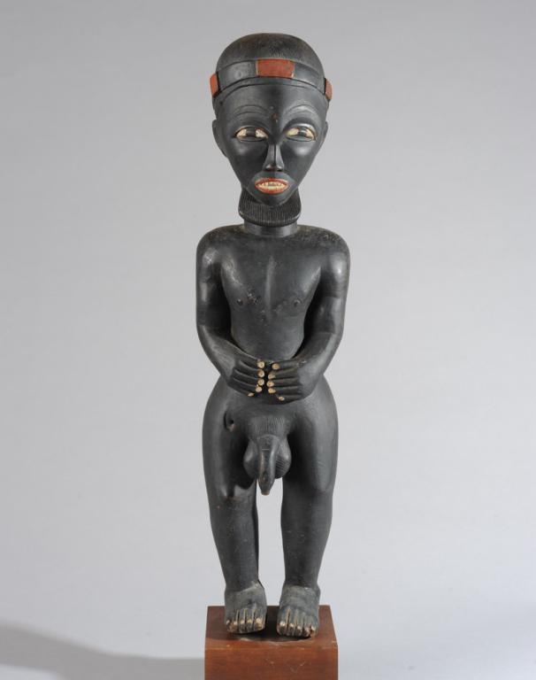 Fon Standing Male Figure: Finely carved hardwood figure of a young man with a headress or crown on his carefully combed hair, a serene and attractive face with rectangular beard, hands resting on his abdomen above a prominent 