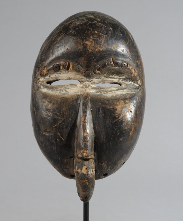 Dan We Bird Mask: Round smooth forehead, narrow eyes whitened with kaolin, slender nose and, in place of the mouth, a bird's beak. Includes custom gallery stand. Mask, approx. 10 1/2" x 6 1/4" x 3 1/2". Provenance: Est