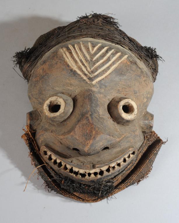 Pende Mask, D. R. Congo: The forehead with incised chevron markings above tube-like eyes, broad nose and wide grimacing smile with many white teeth. Raffia and woven natural fiber headdress, woven raffia beard. Mask approx. 1