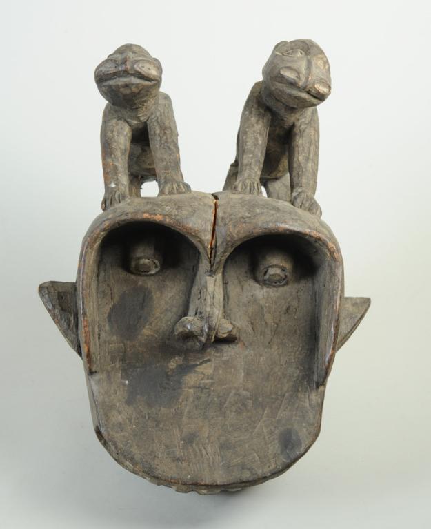Wum Helmet Mask, Cameroon: Gorilla-like face with dramatic, overhanging brow over deep set, protruding eyes, with naturalistic nose and enormous mouth full of long teeth. Two animals with long, flaring nostrils are perched on t