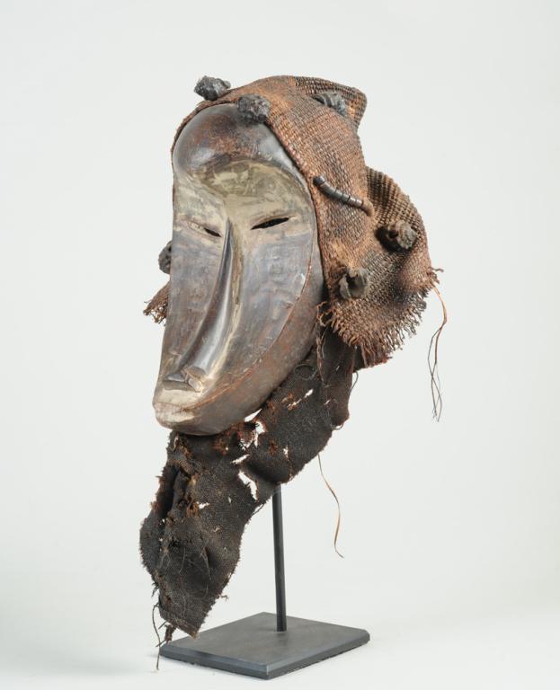Fang Style Mask with Accoutrements: Mask with dramatically scooped facial plane under bulging brow, and with slit eyes, long narrow nose, simplified mouth. Woven grass hood with attached beads and seashells, the chin with remnants of te