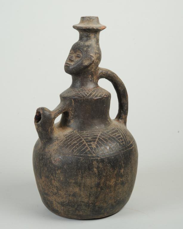 Songye Terracotta Vessel: Black terracotta handled pot with spout, the narrow neck having wide rim and a face with closed, coffee bean eyes under rounded brow. The upper part of the pot with incised geometric patterns. Approx.